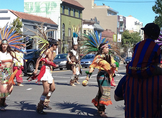 People in colorful costumes and feathers dance in the street.
