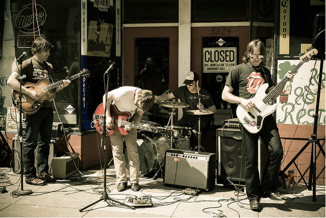 Young men play guitar in front of a cafe.