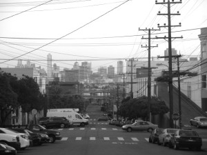 A San Francisco street lined with parked cars and utility poles, leading toward a distant city skyline under an overcast sky.