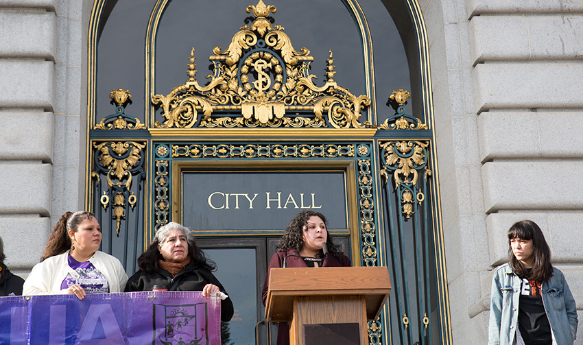 Woman stands at lectern on the steps of city hall. She is flanked by other immigrants' rights activists.
