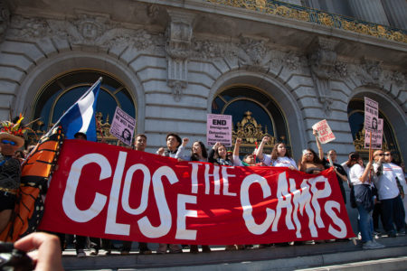 Protesters stand on building steps, holding a large "Close the Detention Camps" banner, with several also clutching smaller signs. In support of immigrants.