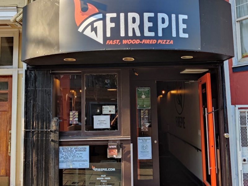 The firepie business store front with door open, various paper fliers posted on front picture window, and a black banner over the door and window with the "Firepie" name and flaming pizza logo in white block letters with orange accents