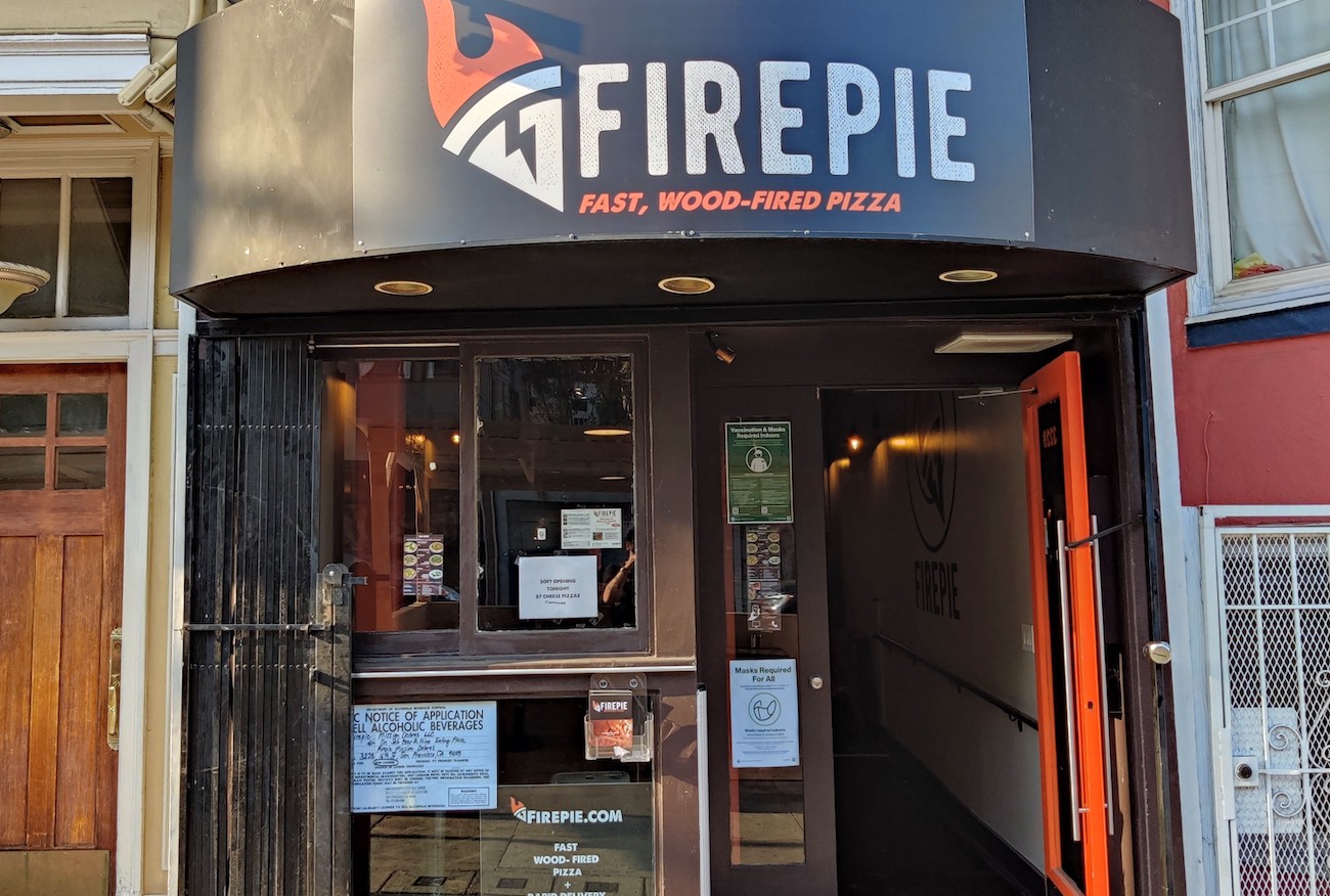 The firepie business store front with door open, various paper fliers posted on front picture window, and a black banner over the door and window with the "Firepie" name and flaming pizza logo in white block letters with orange accents