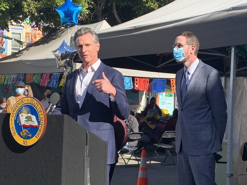 Governor Gavin Newsom is standing at podium with his hand in an emphasizing gesture while standing next to Senator Scott Wiener