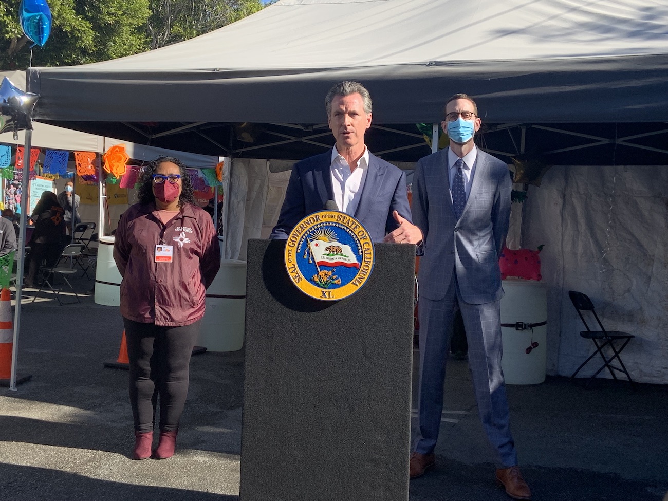 governor gavin newsom stands at a podium addressing press (off camera) while susana rojas stands to his right and senator scott wiener stands to his left in front of a vaccination tent