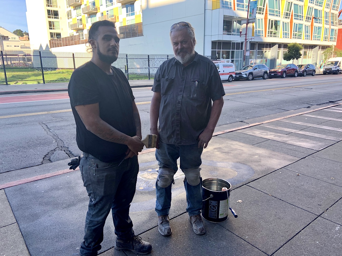 Two men stand on a Mission street sidewalk. They are in work clothes; Jose Medina, left, holds a paintbrush, and Dan Danielson, right, holds a pain roller. They are standing in front of an abstract round shaped terazzo artwork inlaid in the cement with a 5 or 10 gallon bucket of clear sealant.
