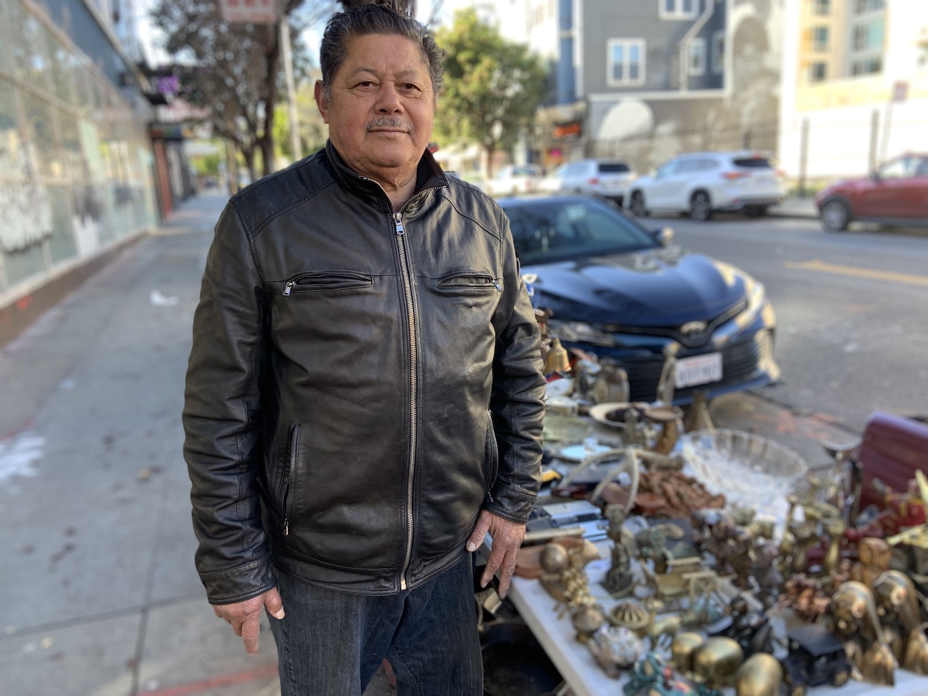 a 69 year old man in a black leather jacket stands next to a table of varied antiques and knick knacks on the sidewalk