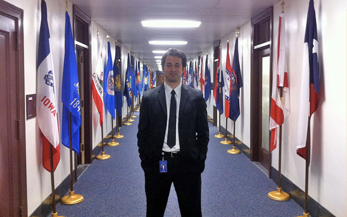 A man stands in a hallway, with flags along the walls behind him.