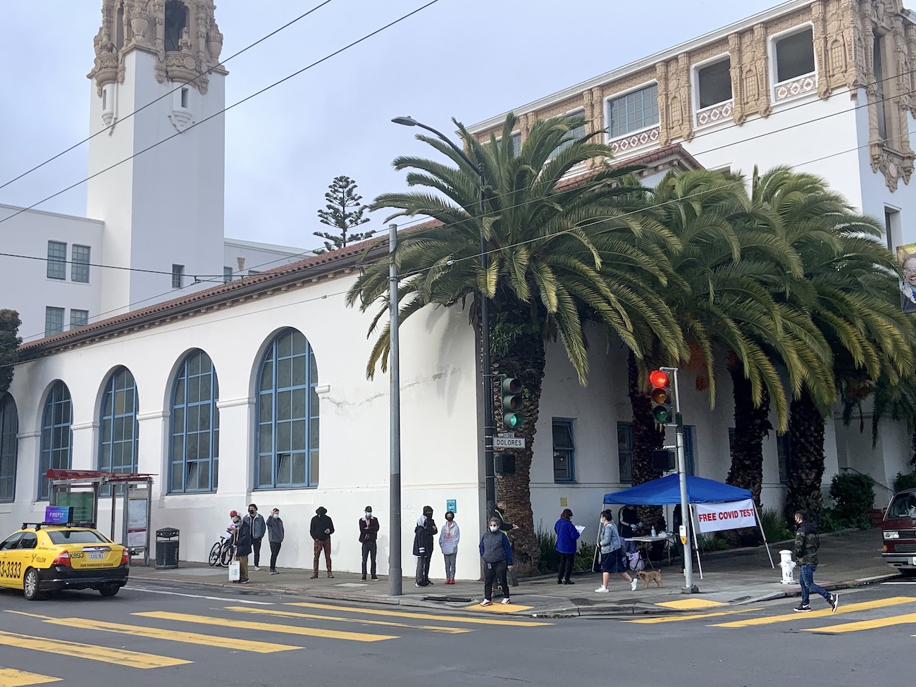 at the corner of 18th and dolores streets, a line of about 15 people forms next to a small white tent with a sign that reads "free covid testing." The line stretches around the Mission High School building which the tent abuts under a row of mature palm trees