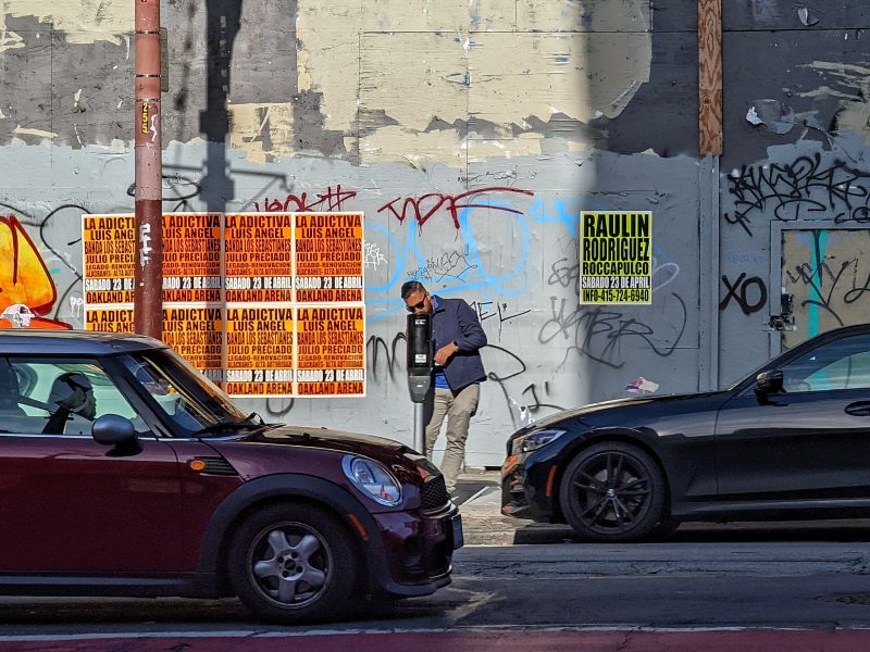 Man putting change into a parking meter on Mission Street. wheat paste in the back.