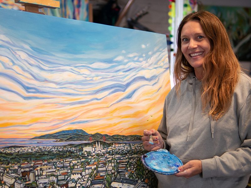 Rachel Znerold smiling in front of a newly painted landscape with a sunset