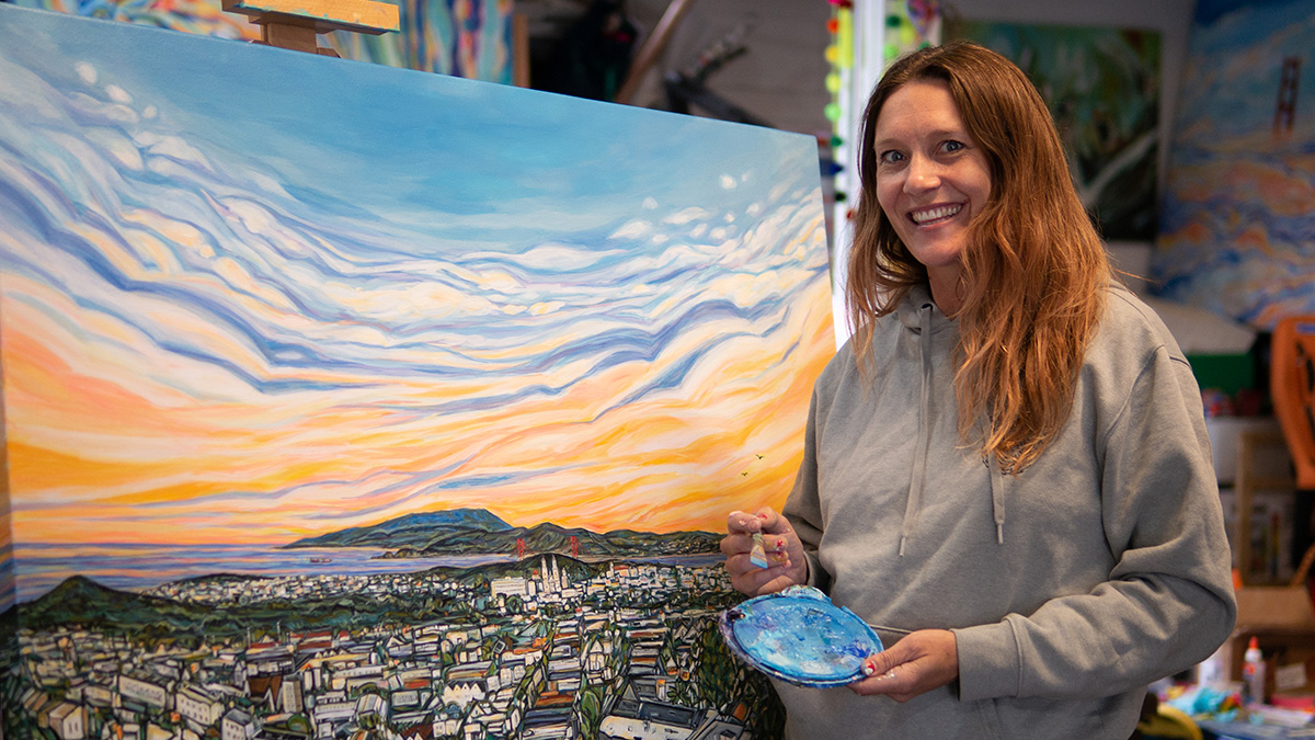 Rachel Znerold smiling in front of a newly painted landscape with a sunset