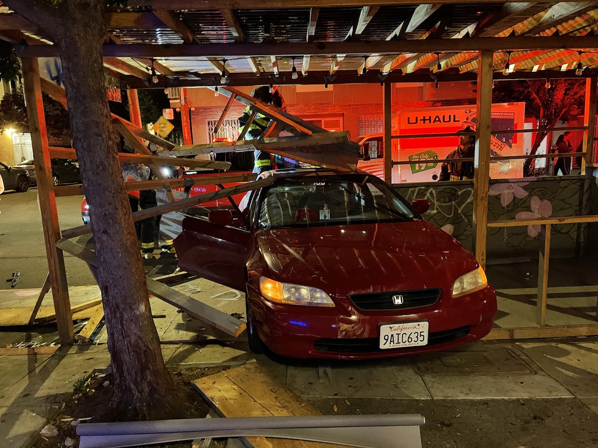 A red Honda is parked in a damaged parklet with plywood hanging onto it.