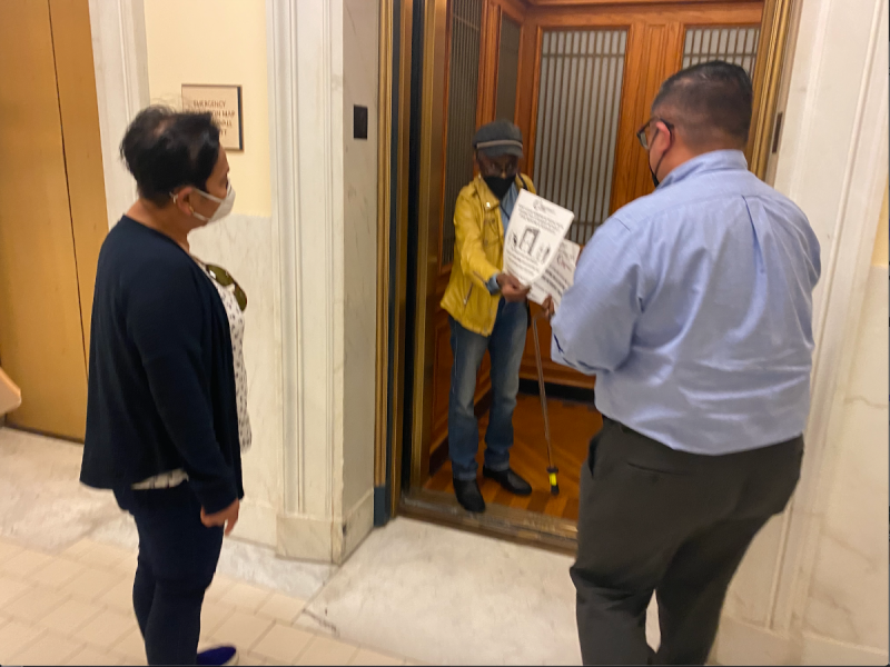 a woman with a cane hands two City Hall workers a flier protesting neglected elevators in SROs