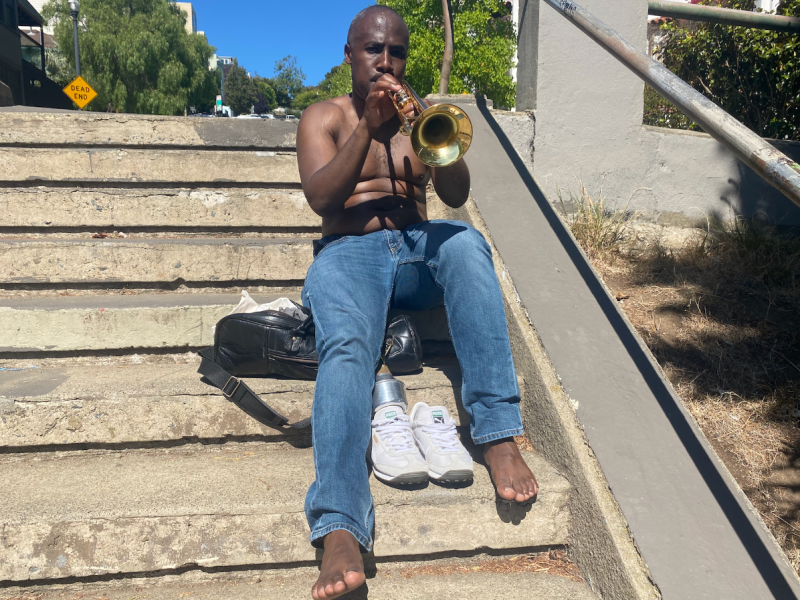 A man sits at the top of the Dolores Park Stairs with his shirt and shoes off. He is playing the trumpet.