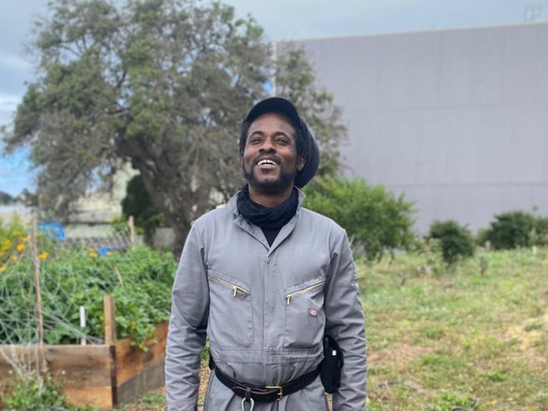 Isaiah Powell, wearing grey coveralls and a black rasta hat, stands in the middle of his garden smiling and looking up at the sky