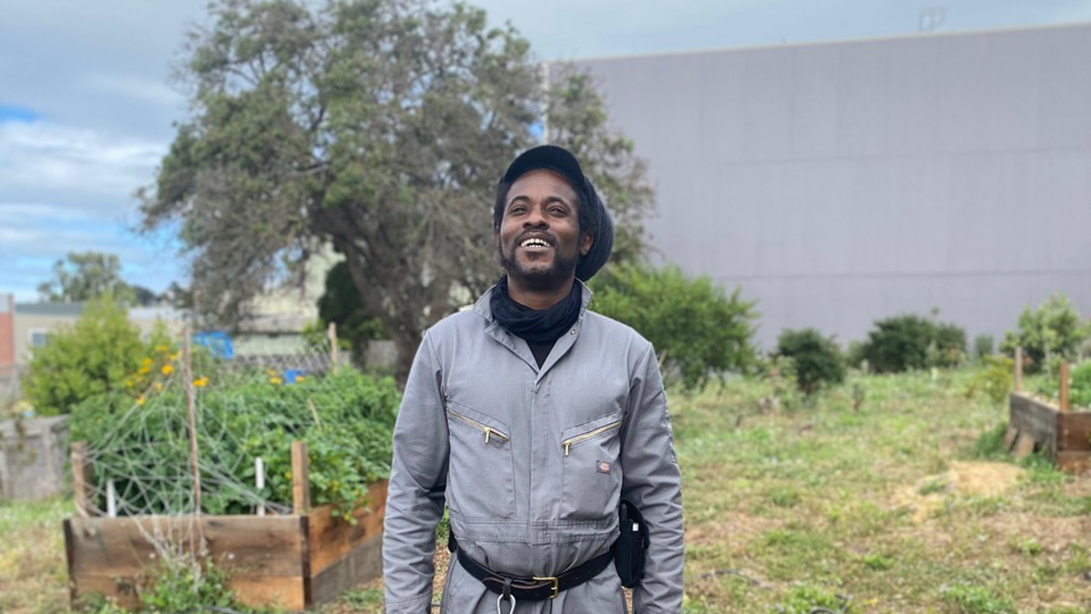 Isaiah Powell, wearing grey coveralls and a black rasta hat, stands in the middle of his garden smiling and looking up at the sky