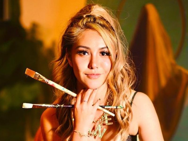 Photo of Yijun Ge, holding two long paintbrushes in her hand holding her chin with a gold and green background