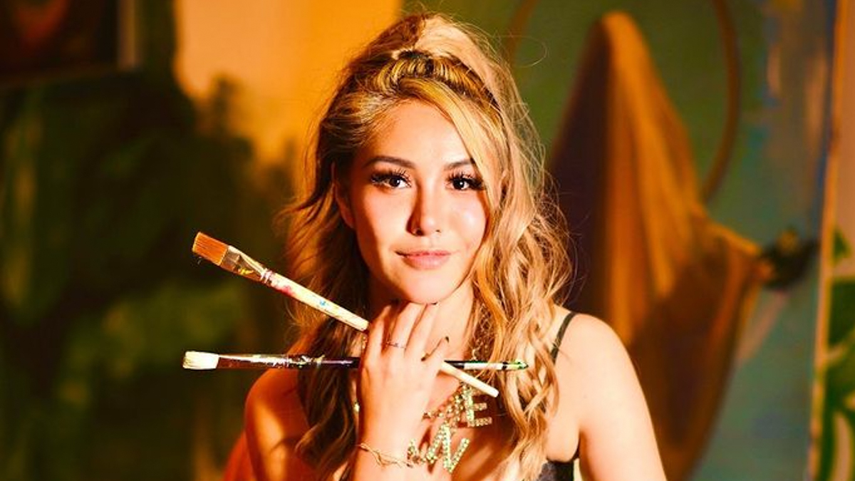 Photo of Yijun Ge, holding two long paintbrushes in her hand holding her chin with a gold and green background