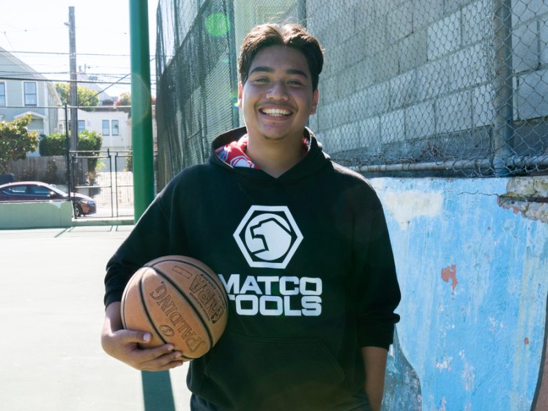 Photo of Alex Romero. Holding a basketball in his right hand, he smiles wearing a black hoodie standing in front of a blue mural at an outdoor basketball court.