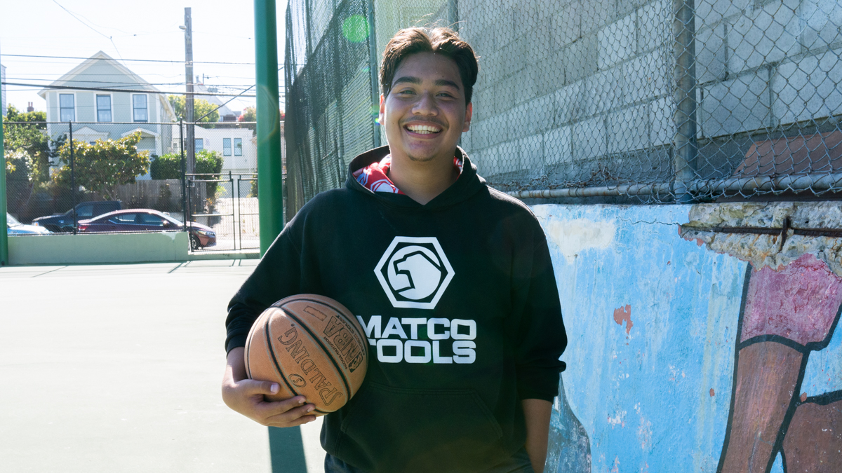 Photo of Alex Romero. Holding a basketball in his right hand, he smiles wearing a black hoodie standing in front of a blue mural at an outdoor basketball court.