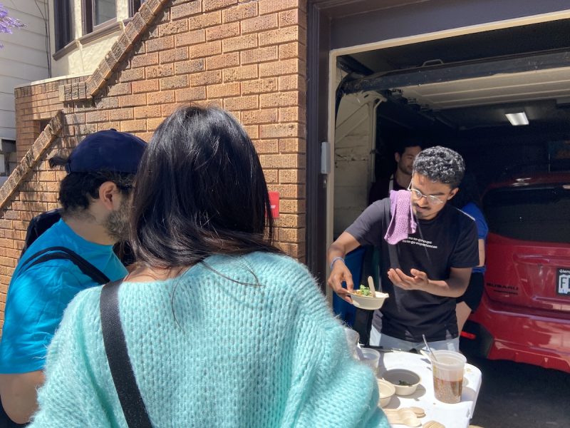 A man in glasses and a bowl serves a bowl of chili crisp to two folk outside his garage.