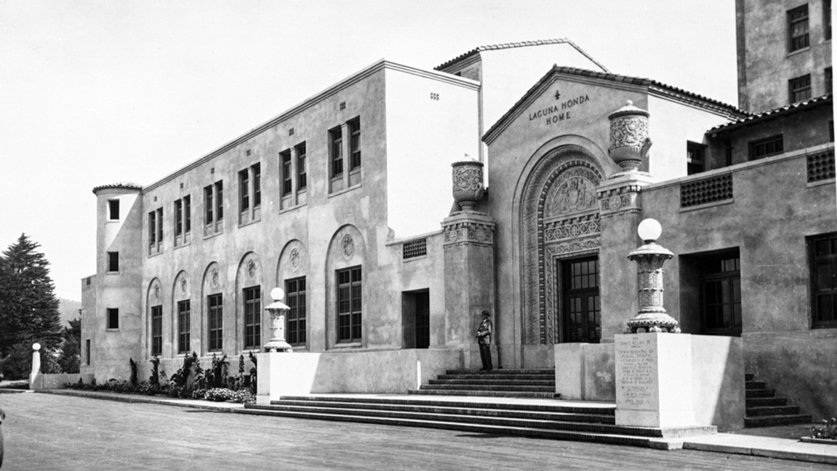 A stone-built building stands backlit against the sun. Words across the top read, Laguna Honda Hospital