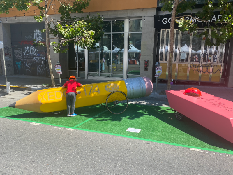 A young boy leans over a pencil derby car. A giant eraser car sits next to him.