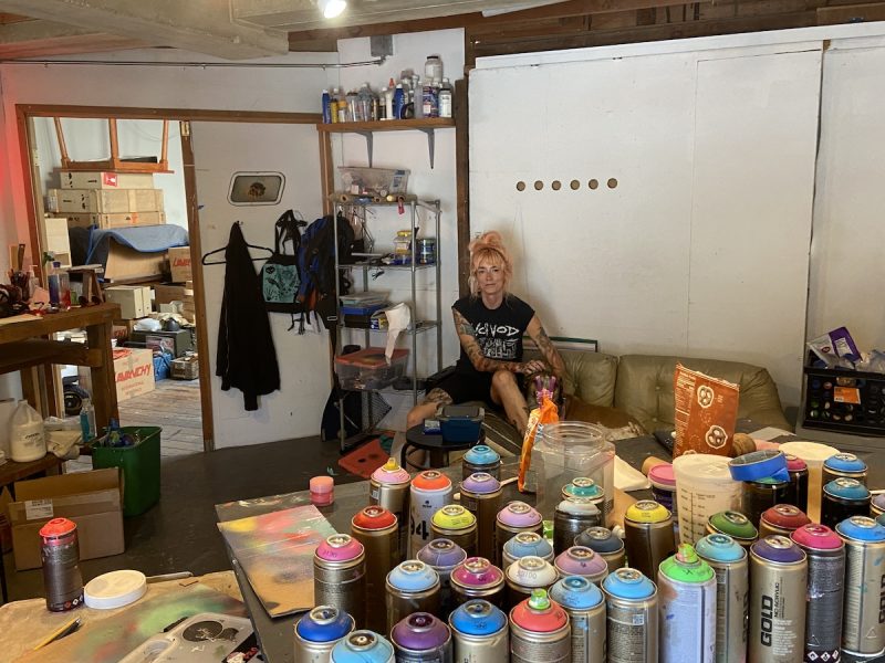 An artist with pastel pink hair and tattoos on her arms is seated on the couch with her dog, Hank. Around her is spray paint cans of every color; she is a spray paint artist.