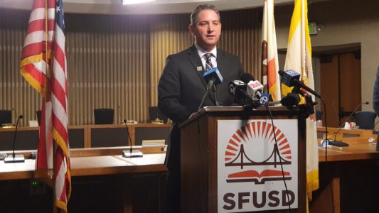 A man in a suit speaks at a podium labeled "SFUSD," surrounded by microphones. American flags are in the background.