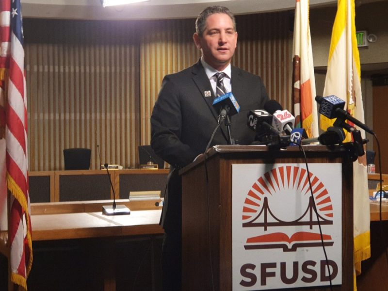 A man in a suit speaks at a podium labeled "SFUSD," surrounded by microphones. American flags are in the background.