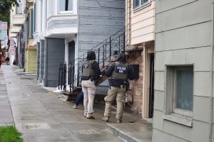 Two people in Police HSI vests stand in front of a house