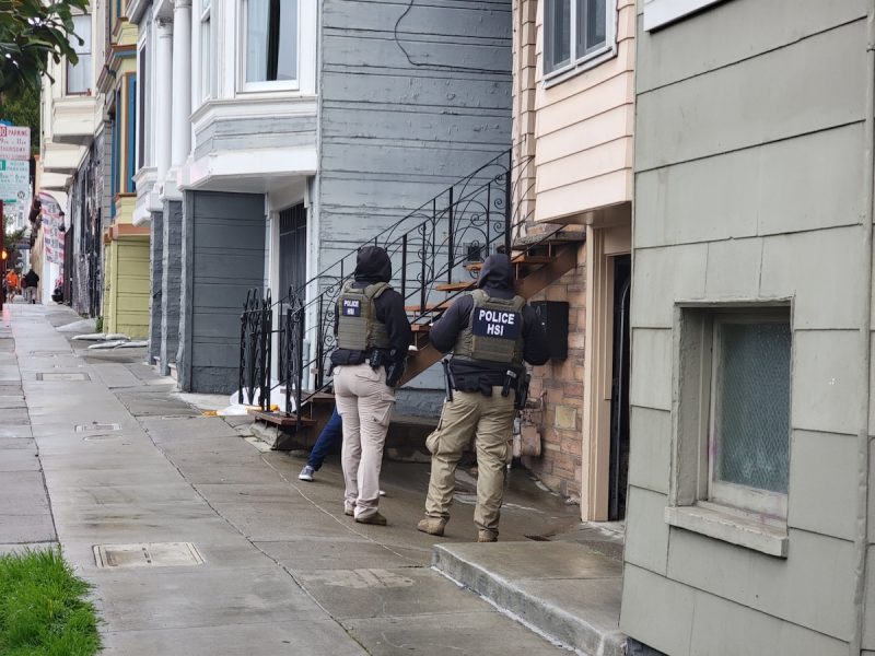 Two people in Police HSI vests stand in front of a house