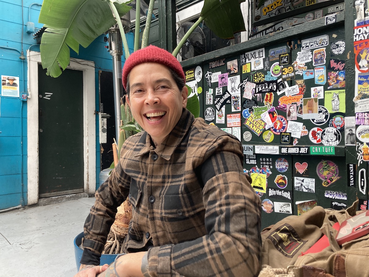 A woman in a beanie and a flannel shirt smiles in front of a wall of stickers