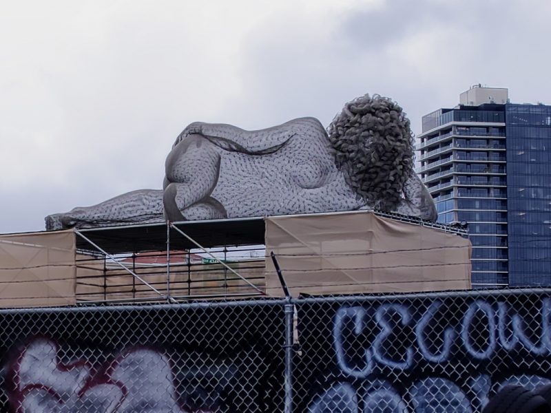 A wireframe sculpture woman with her back to us, lying on a roof looking toward downtown.