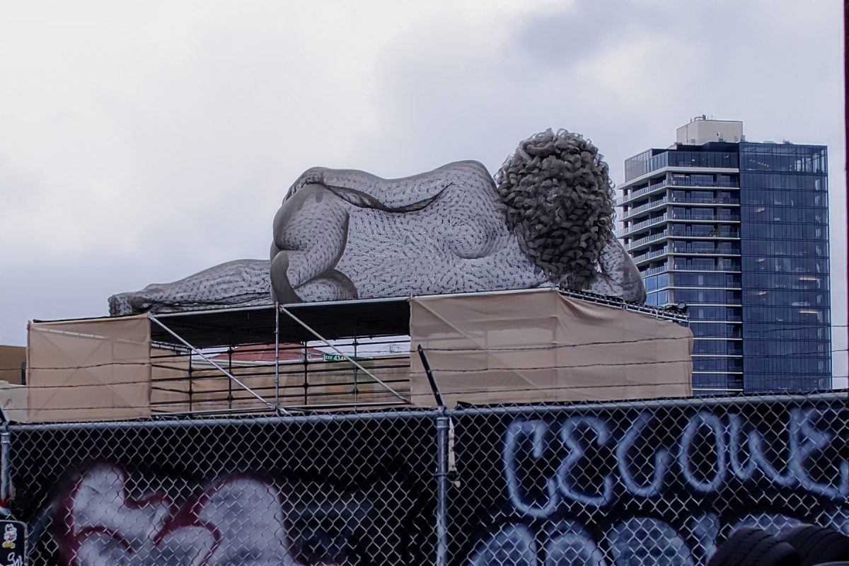 A wireframe sculpture woman with her back to us, lying on a roof looking toward downtown.