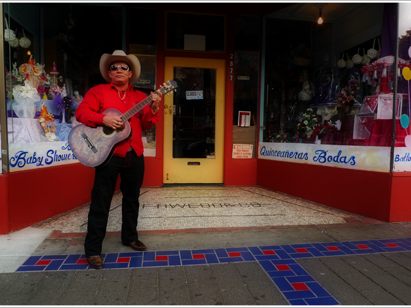 A man wearing a cowboy hat and standing in front of a store with his guitar