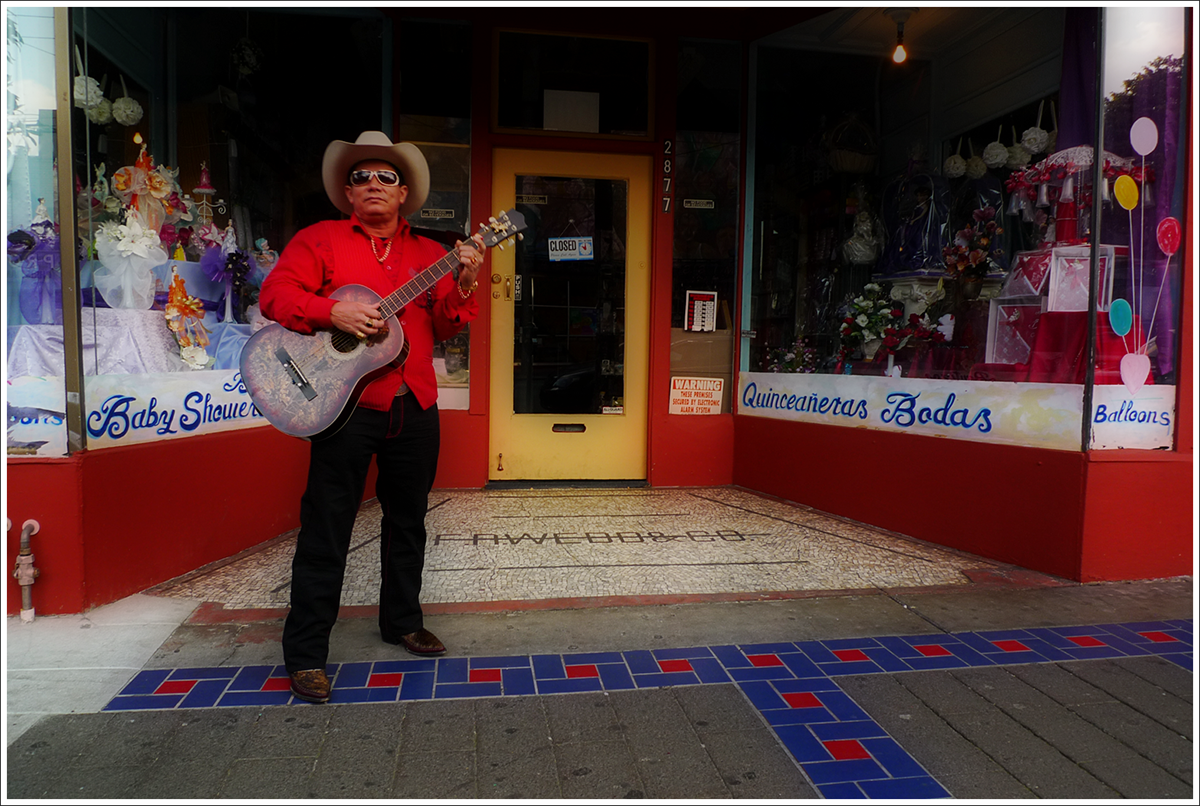 A man wearing a cowboy hat and standing in front of a store with his guitar