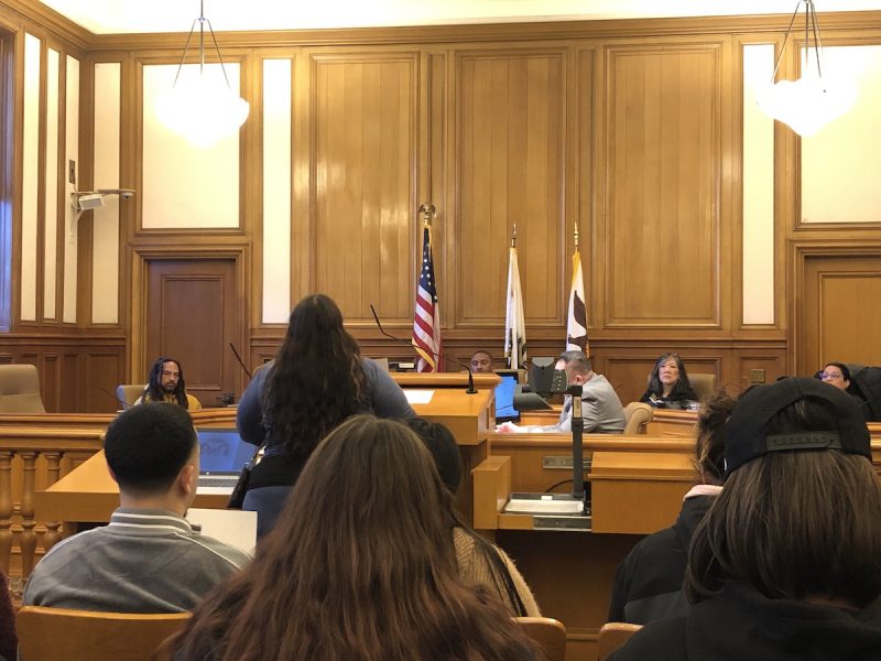 a woman standing in front of a podium, facing the sheriff's oversight board, in a hearing room at the city hall