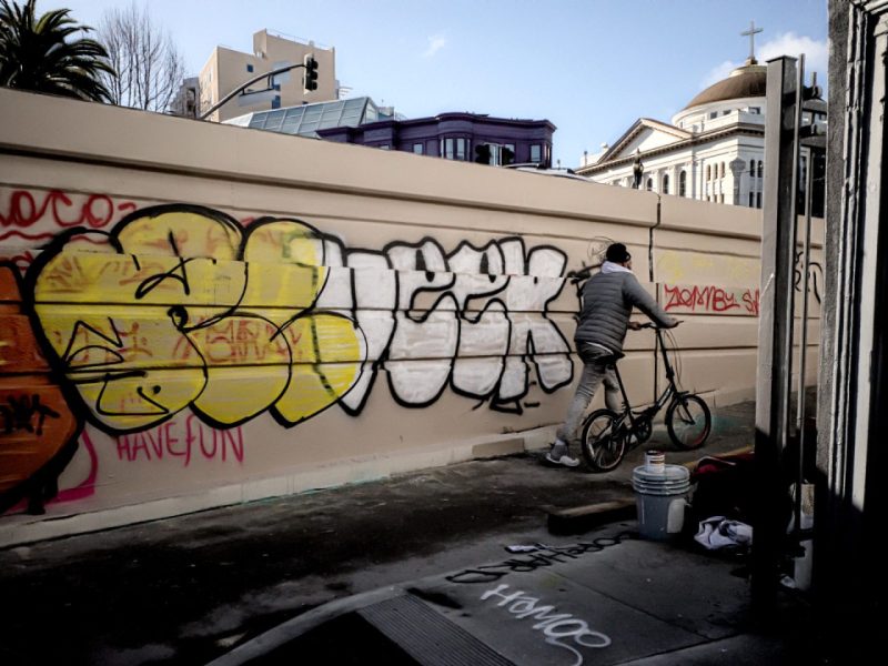 A wall full of large graffiti letters, red, yellow and white and a man on a bike leaving the alleyway.