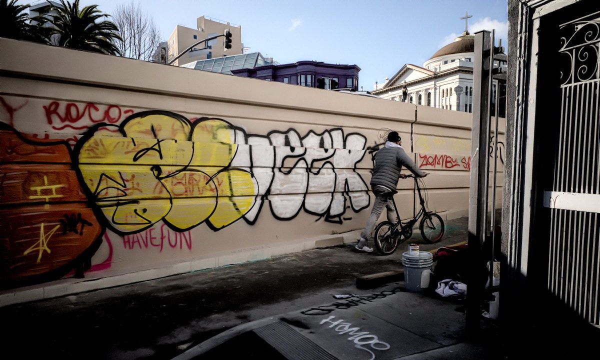 A wall full of large graffiti letters, red, yellow and white and a man on a bike leaving the alleyway.