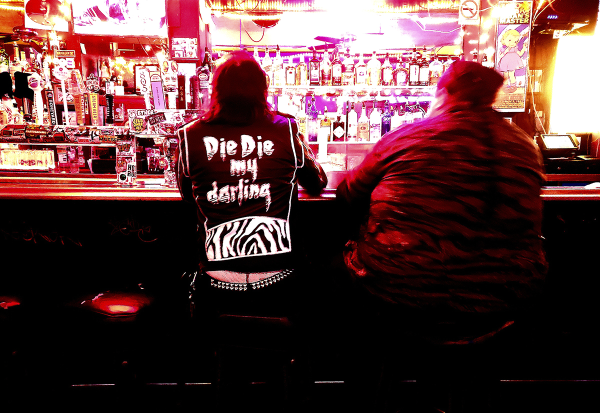 The backs of two men sitting a bar. One has a black hoodie with the words, die die my darling on the back.