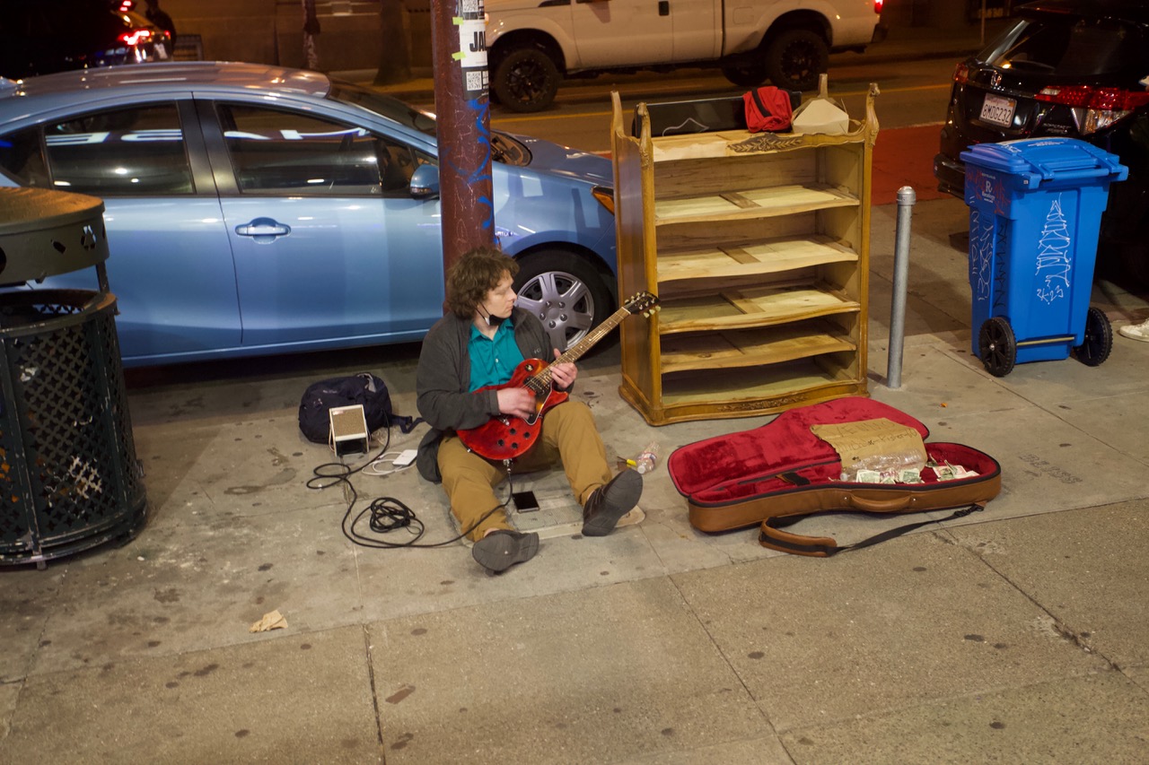 A man sitting on the sidewalk, his legs out in front of him, guitar in his hand. An empty book case is beside him.