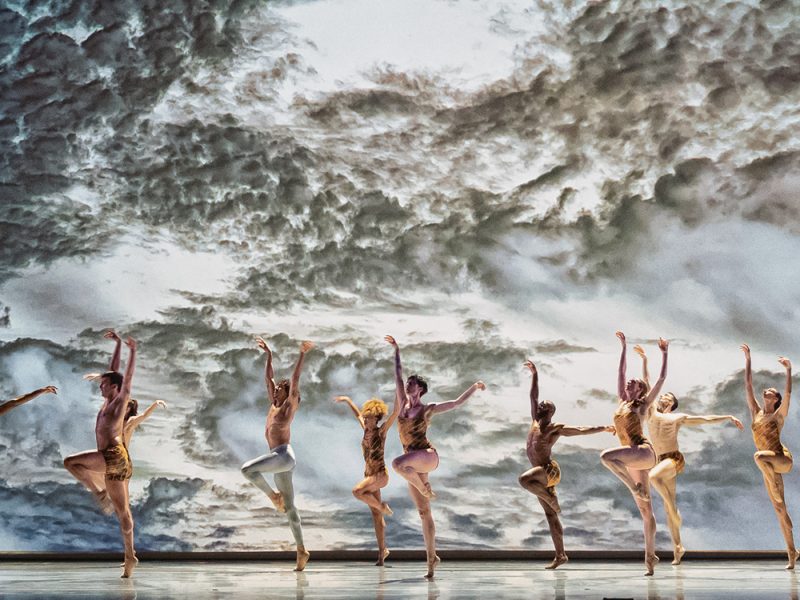 A group of dancers across a stage, a background of sea.