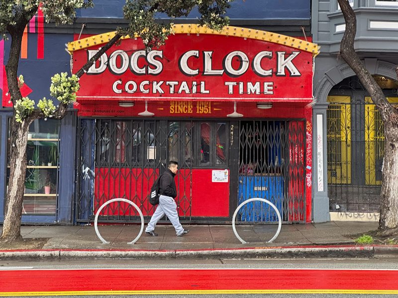A red front of a cocktail place called Doc's Clock. A man is walking by.