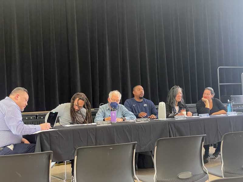 A line of members of a board sitting behind a long table with a black curtain in the back.