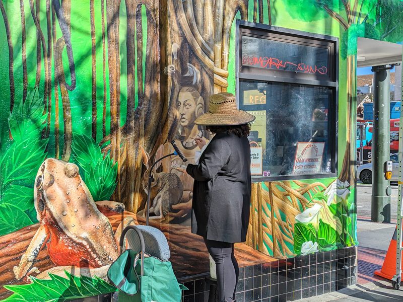 A woman to her back with us painting a mural with lots of green in it.