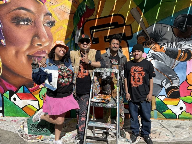 A muralist in a skirt and bucket hat throws up a peace sign, three other muralists stand behind a ladder, and all stand in front of their newly painted Giants mural.