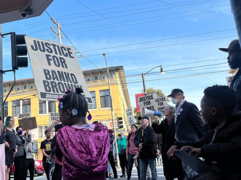Little girl holding a sign saying "Justice for Banko Brown" in foreground, with crowd in background