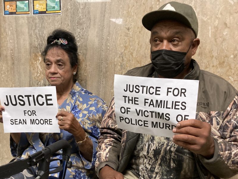A woman and man hold signs that read "Justice for Sean Moore" and "Justice for the families of victims of police murder"